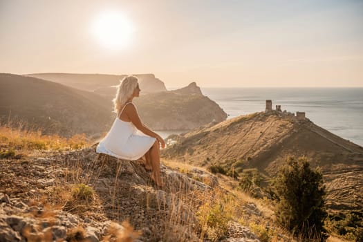 A woman is sitting on a hillside overlooking the ocean. She is wearing a white dress and has blonde hair. The scene is serene and peaceful, with the ocean in the background