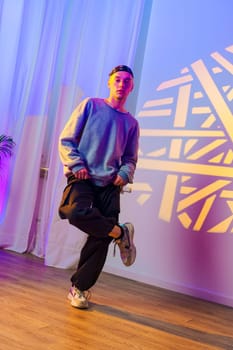 A young man, wearing a blue sweater and black pants, stands with one leg bent, his foot raised. He is in a dance studio, and a colorful light pattern is projected onto the wall behind him.