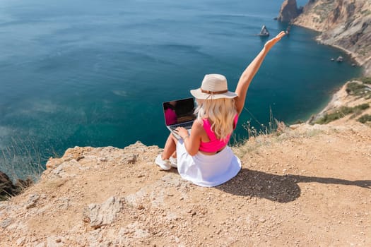 A woman is sitting on a rocky beach with a laptop in front of her. She is wearing a pink top and a straw hat. She is waving her hand in the air, possibly to celebrate or to show excitement