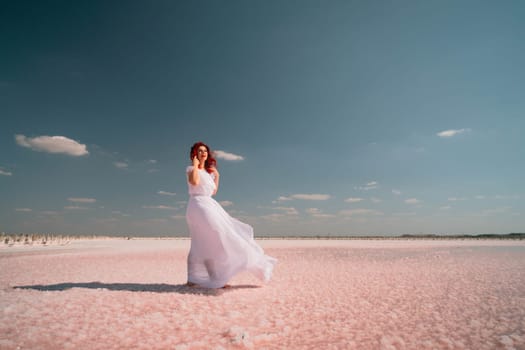 A woman in a white dress stands on a pink salt flat, her dress billowing in the wind. The sky is blue and cloudless.