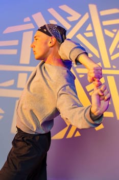 A young man wearing a gray sweatshirt, black pants, and a bandana dances in front of a wall. The wall has geometric patterns in white projected onto it. The lighting is warm and vibrant.