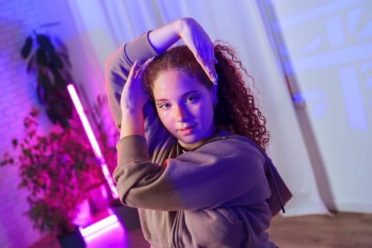 A young woman with curly hair and a grey hoodie is posing in a room with white walls and a pink neon light behind her. She is illuminated by a soft blue light.