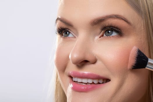 A smiling model applies blush with a brush, showcasing beauty and makeup artistry in a close-up shot.