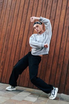 A man wearing a grey sweatshirt and black pants is dancing in front of a wooden wall. His right leg is extended forward as he leans back. He is wearing a black bandana around his head.