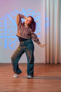 A young woman with curly hair dances in a studio. She is wearing a brown jacket, blue jeans, and white sneakers. The walls are white and there is a white curtain in the background.