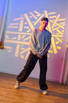 A person wearing black pants, a gray sweatshirt, and a bandana stands in a studio. The person is posing with their arms crossed and looking at the camera.