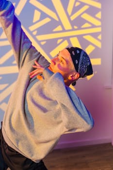 A young person wearing a gray sweatshirt and bandana dances against a wall with abstract lighting. They are caught mid-pose with their arm extended overhead and their body contorted in a dynamic way.