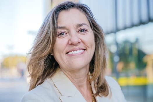 Close-up portrait of a cheerful middle-aged woman in a beige blazer, smiling warmly in an outdoor urban setting with a blurred modern background.