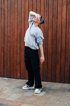 A person stands in front of a wooden wall with their hand over their eyes. They are wearing a grey sweatshirt, black pants, a bandana, and white sneakers.