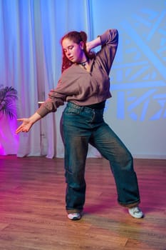 A young woman with long, curly red hair is dancing in a studio. She is wearing a brown zip-up jacket, blue jeans, and white sneakers.