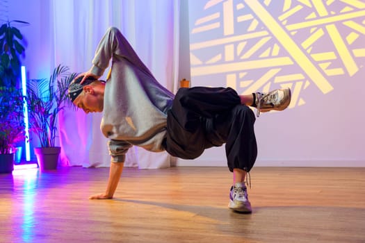 A breakdancer in a gray sweatshirt and black pants performs a move on a wooden floor.