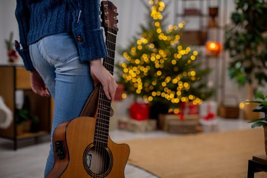 An individual is holding a lovely acoustic guitar while standing next to a beautifully adorned Christmas tree