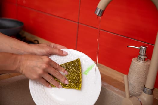 A hand is carefully washing a plate using a sponge under the soothing flow of running water in a kitchen sink