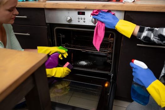 Two individuals are actively engaged in cleaning an oven using colorful gloves and various supplies in a bright kitchen