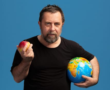 A man stands confidently against a solid blue background, holding an apple in one hand and a colorful globe in the other. His expression conveys deep thought about nutrition and the world.