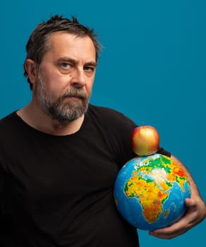 A man with a beard holds a colorful globe of the Earth in one arm and a bright red apple in the other. His serious expression highlights the connection between nature and the planets health.