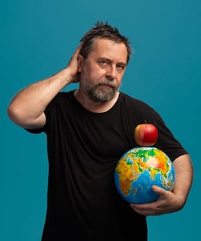A man with a beard poses thoughtfully while cradling a globe in one arm and balancing a bright red apple on top. Set against a vibrant blue backdrop, he captures a moment of reflection.