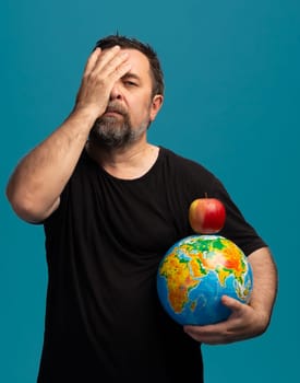 A man stands against a vibrant blue backdrop, one hand covering his face while holding a globe in the other. An apple perches atop the globe, symbolizing a blend of worry and hope for the world.