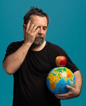 A man with a beard stands thoughtfully, holding a globe in one hand while an apple rests on top. His expression reveals contemplation, set against a vibrant blue background.