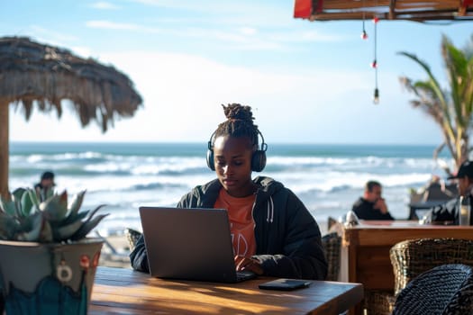 A woman is sitting at a table with a laptop, wearing headphones and looking at the screen. She is in a beach setting, with a potted plant nearby. Concept of relaxation and leisure