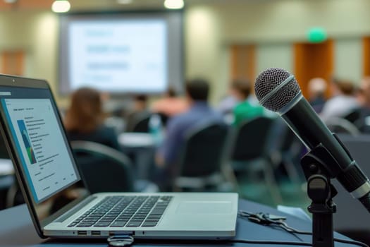 A microphone is on a table next to a laptop. The microphone is black and silver. The laptop is open and has a screen. The scene appears to be a presentation or a lecture