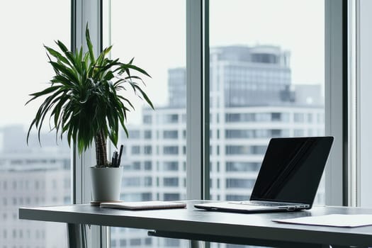 A laptop is on a desk next to a potted plant. The plant is a large green plant with a white pot. The laptop is open and the screen is black. The desk is in a window with a view of the city