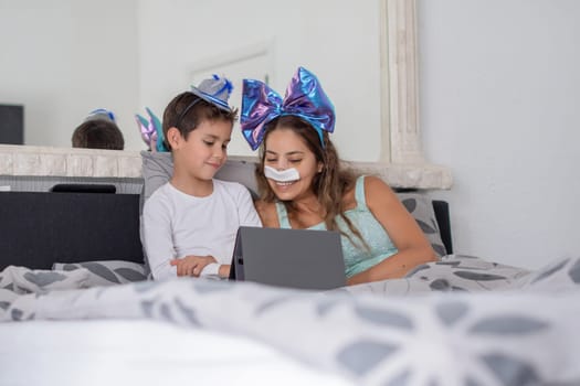 Smiling mother with bandage on nose lies in cozy bed and plays on tablet with son,mother has big blue bow on head and son has blue hat, carnival in bed to lift mood, relaxing,