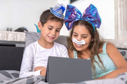 Smiling mother with bandage on nose lies in cozy bed and plays on tablet with son,mother has big blue bow on head and son has blue hat, carnival in bed to lift mood, relaxing,