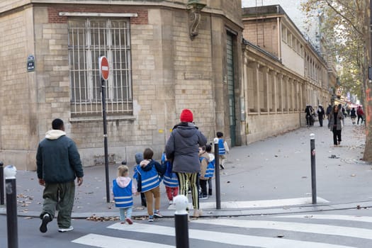 A caregiver expertly guides a group of children dressed in blue safety vests down a bustling Parisian street. Paris, France - October 24, 2024