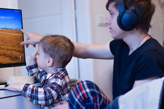 Two children are sitting at the computer. The kid sits on the teenager's arms, and the older brother shows him a picture on the monitor. High quality photo