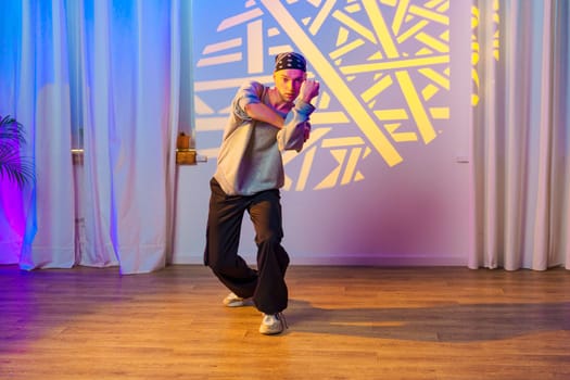 A young man wearing a bandana and loose-fitting clothing dances in a studio. He is surrounded by colorful lighting, casting an abstract pattern on the wall behind him.