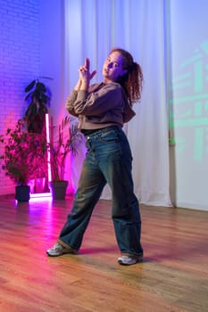 A young woman with curly red hair, wearing a brown sweater and blue jeans, strikes a pose while dancing in a studio.