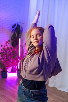 A young woman wearing a grey hoodie and blue jeans is stretching in a studio illuminated by colorful lights. She has red hair and is standing with her arms above her head.
