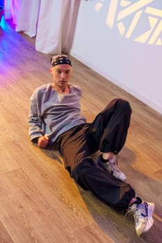 A young man, wearing a grey sweatshirt, black pants, and a bandana, is lying on a wood floor in a dance studio. The man's legs are outstretched, and he is looking directly at the camera.
