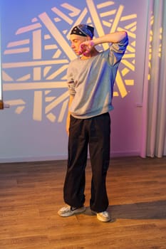 A young man in a grey sweatshirt, black pants, and white sneakers stands in a room. A geometric pattern of light is projected onto the wall behind him.