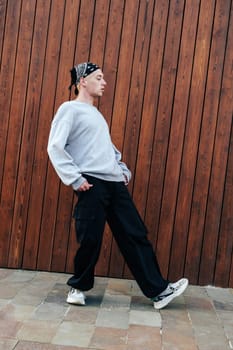 A young man wearing a grey sweatshirt, black pants, and a bandana walks past a wooden wall. The man's feet are slightly turned inward, and his hands are in his pockets.