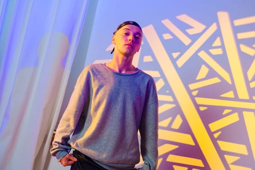 A young man is standing in front of a wall with yellow and purple geometric patterns. He is wearing a gray sweater and black pants. He is looking directly at the camera with a neutral expression.