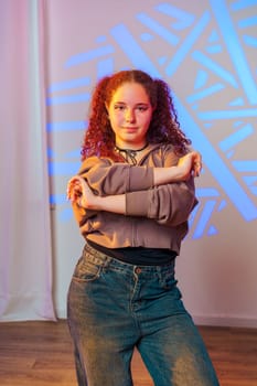 A young woman stands in a studio with her arms crossed, facing the camera. She has curly red hair and is wearing a light brown hoodie and blue jeans.