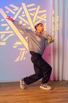 A young man with a grey sweatshirt, black pants, and a bandanna is dancing in front of a wall with a geometric pattern projected onto it.