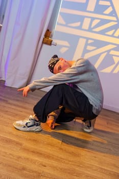 A young male dancer wearing a gray sweatshirt and black pants is in a low squat pose, looking towards the camera. The dancer's left hand is on the ground, while the right arm is outstretched.