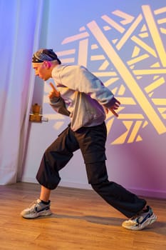A young man in a grey sweatshirt, black pants, and a bandana dances in a studio. The wall behind him is lit with a geometric light projection.