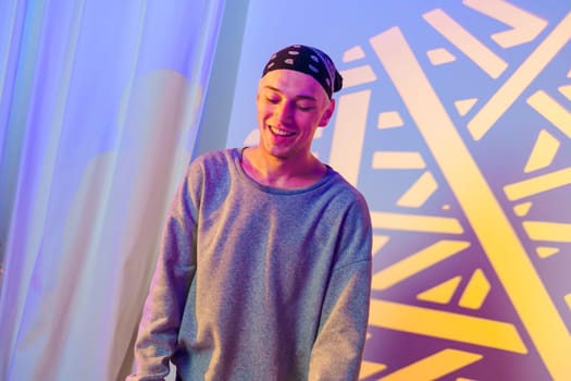 A young man with a bandana smiles as he stands in front of a blue and yellow patterned backdrop. The background is blurred, and the man is in focus. He wears a grey sweatshirt.