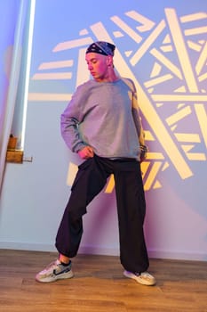 A young man wearing a black bandana, a grey sweatshirt, black cargo pants, and white sneakers strikes a pose in a dance studio. There is a colorful backdrop behind him.