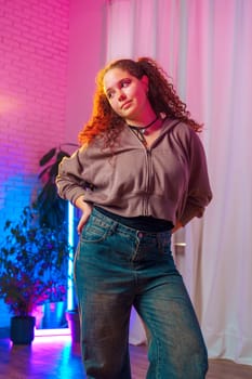 A young woman with curly red hair tied up in a ponytail, wearing a grey zippered jacket, a black shirt, and blue jeans, is standing in a room with pink and blue lighting.