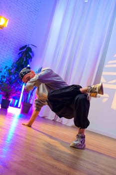 A male dancer wearing a grey sweater, black pants, and a black bandana is performing a breakdancing move in a studio. The floor is wooden and there are plants in pots in the background.