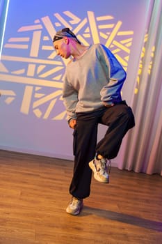 A young person in a grey sweater, black cargo pants, and sneakers dances with one leg lifted in front of a white wall lit by a colorful geometric projection.