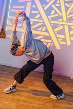 A dancer wearing a gray sweatshirt and black pants is stretching in a studio with a large geometric light projection on the wall. The dancer is bending over, reaching their arm towards the ceiling.