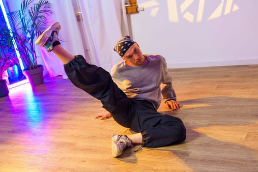 A dancer wearing black pants, a gray sweatshirt, and a bandana is in a dance studio. Their right leg is extended forward, and their left hand is on the floor.