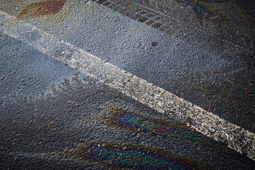 Multicolored oil spill generating a rainbow sheen on the dark pavement, complemented by a white traffic line