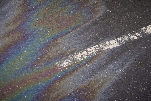 Colorful gasoline slick creating a shimmering rainbow effect on the dark asphalt, alongside a white road marking
